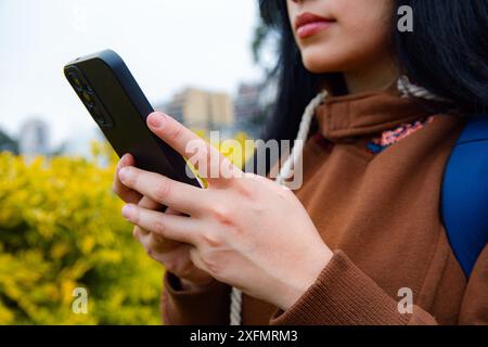 Nahaufnahme einer nicht erkennbaren Frau, die ihr Handy mit den Händen hält, während sie es benutzt, sie ist im Park, sie trägt braune Kleidung, Technologiekonzept, Co Stockfoto