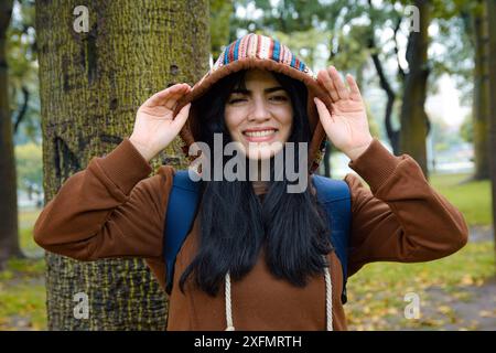 Porträt der lustigen Latin Jungen Frau im Park Sie steht neben einem Baum, trägt einen braunen Pullover und hält einen Hoodie mit den Händen, während sie lächelt Stockfoto