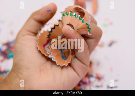 Eine Hand hält einen Haufen bunter Bleistiftspäne, die wahrscheinlich gerade von einem Bleistiftspitzer entfernt wurden. Auf einer weißen Oberfläche im Backgr befinden sich weitere Späne Stockfoto