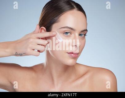 Frau, Porträt und Anwendung von Creme im Studio, Hydration und glatte Schönheit auf blauem Hintergrund. Weibliche Person, Kosmetik und Gesichtslotion Stockfoto