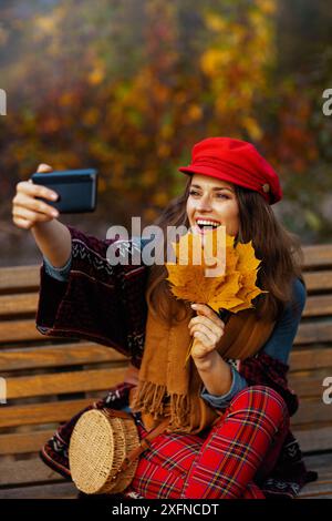 Hallo Herbst. Glückliche, elegante 40-jährige Frau mit rotem Hut mit Herbstblättern, Schal und Tasche, die auf einer Bank in der Stadt sitzt Stockfoto