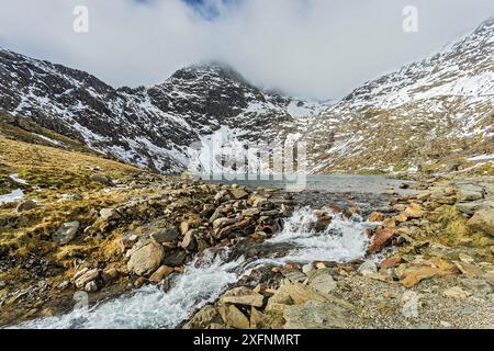 Lake Glaslyn mit dem Abfluss der Quelle des Afon (River) Glaslyn mit dem Gipfel des Mount Snowdon bedeckt mit Wolken im Hintergrund, Snowdonia National Park, North Wales, UK, März. Stockfoto