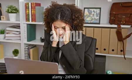 Eine junge hispanische Frau mit lockigem Haar, die Stress am Arbeitsplatz hat, Kopfhörer und Anzug trägt. Stockfoto