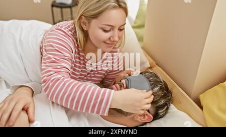 Eine sanfte Frau legt in einem gemütlichen Schlafzimmer eine Schlafmaske auf das Gesicht eines Mannes, die Intimität und Sorgfalt zwischen einem Paar darstellt. Stockfoto