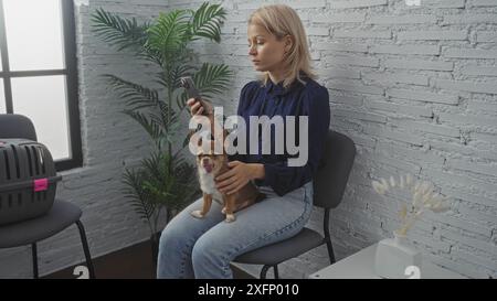 Junge blonde Frau mit chihuahua-Hund im Wartezimmer einer Tierklinik, die ihr Telefon hält. Stockfoto