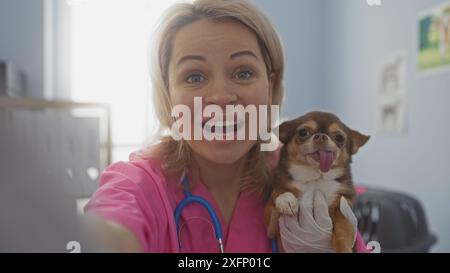 Eine junge kaukasische Tierärztin macht ein fröhliches Selfie mit einem blonden chihuahua-Hund in einem Tierklinikzimmer. Stockfoto