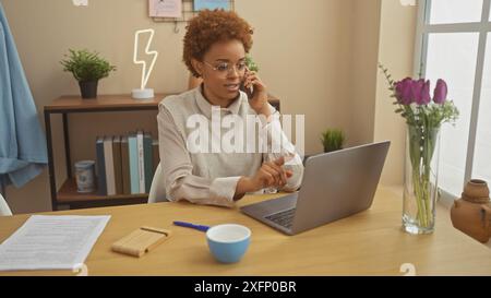Eine afroamerikanische Frau arbeitet von zu Hause aus und verwaltet Aufgaben auf ihrem Laptop, während sie in einer gemütlichen Inneneinrichtung am Telefon telefoniert. Stockfoto