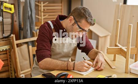 Ein bärtiger Mann skizziert Entwürfe in einer hellen Tischlerei, umgeben von Holzmöbeln und Werkzeugen. Stockfoto