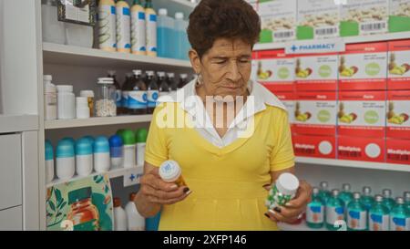 Eine hispanische ältere Frau mit kurzen Haaren und gelbem Kleid untersucht Arzneiflaschen in einem gut sortierten Apotheken-Innenraum. Stockfoto