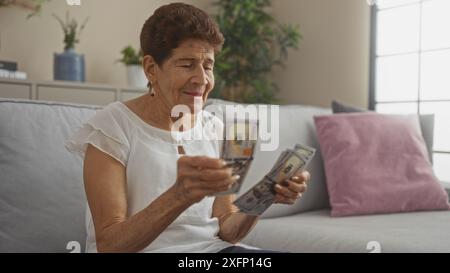 Eine ältere hispanische Frau mit kurzen Haaren zählt einen Stapel von uns Dollarscheine, während sie auf einer Couch in einem gemütlichen Wohnzimmer sitzt. Stockfoto