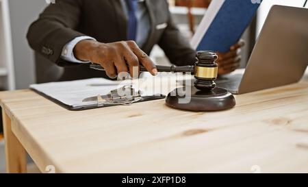 afroamerikanischer Mann im Anzug, der einen Hammer am Schreibtisch mit Laptop im Büro benutzt und Recht, Autorität und Professionalität vermittelt. Stockfoto