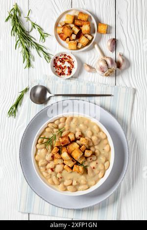rosmarin Knoblauch weiße Bohnensuppe mit hausgemachten Croutons in einer weißen Schüssel mit Löffel und Einlagen auf weißem Holztisch, vertikale Ansicht von oben Stockfoto