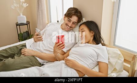 Ein interrassisches Paar entspannt sich in seinem Schlafzimmer und genießt die gemeinsame Zeit, während es ein Smartphone benutzt. Stockfoto