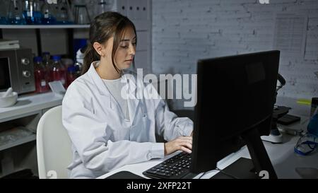 Eine fokussierte hispanische Frau arbeitet fleißig an einem Computer in einem Labor, umgeben von wissenschaftlichen Geräten. Stockfoto