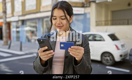 Die junge hispanische Frau checkt ihr Handy und ihre Kreditkarte in einer sonnigen Stadtstraße. Stockfoto