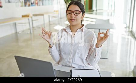 Eine junge hispanische Frau meditiert an ihrem Arbeitsplatz und fördert Wohlbefinden und Achtsamkeit. Stockfoto