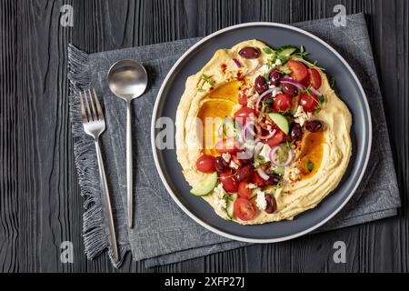 Beladen griechischen Hummus mit frischen Gurken, Oliven, Tomaten, Fetakäse, roten Zwiebeln und Thymian auf Teller auf schwarzem Holztisch mit Löffel und Gabel, flach Stockfoto