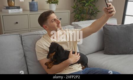 Ein junger hispanischer Mann, der in seinem Wohnzimmer sitzt, macht ein Selfie, während er seinen Hausdackel bequem auf der Couch hält. Stockfoto