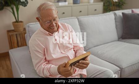 Ein älterer Mann mit grauen Haaren sitzt in einem gemütlichen Wohnzimmer, lächelt warm auf ein gerahmtes Foto in seinen Händen, gönnt sich einem Moment der Nostalgie und des Glücks Stockfoto