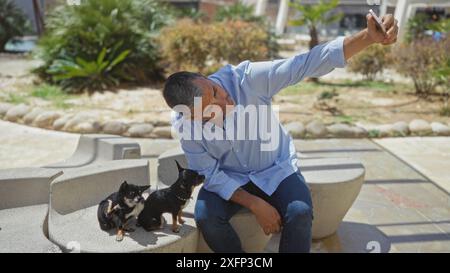 Ein erwachsener hispanischer Mann macht ein Selfie mit zwei kleinen Chihuahuas in einem Outdoor-Stadtpark. Stockfoto
