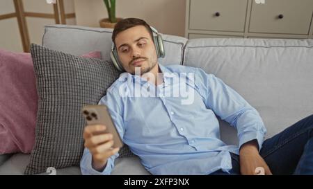 Hübscher hispanischer Mann mit Bart und Kopfhörern und blauem Hemd, entspannend auf einer Couch im Wohnzimmer und auf sein Handy in einem Innenheim Stockfoto