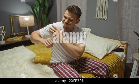 Ein hübscher, junger hispanischer Mann mit Bart sitzt auf seinem Bett im Schlafzimmer, trägt Pyjamas und massiert seine Schulter, zeigt ein zwangloses Zuhause Stockfoto