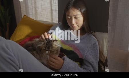 Eine junge asiatische Frau ruht sich bequem in ihrem Schlafzimmer aus und streichelt sanft eine flauschige Katze auf ihrem Bett. Stockfoto