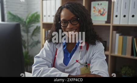 Eine lächelnde afroamerikanische Ärztin mit lockigen Haaren sitzt drinnen in einem Krankenhausarbeitsplatz, trägt einen Labormantel und Stethoskop und strahlt Conf aus Stockfoto