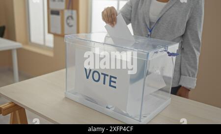 Eine junge afroamerikanische Frau gibt ihre Stimme in einem Wahlraum ab und legt eine Stimmzettel in ein klares Feld mit der Aufschrift „Stimme“, in dem sie ihre Participatio darstellt Stockfoto