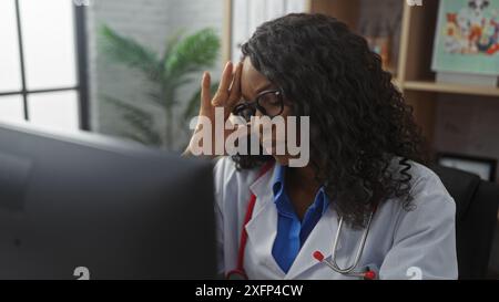 Eine junge afroamerikanische Ärztin mit lockigem Haar und Brille, die Kopfschmerzen an ihrem Kliniktisch drinnen hat. Stockfoto