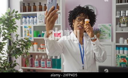 afroamerikanische Apothekerin mit lockigen Haaren, die ein Selfie macht, während sie eine Pille-Flasche in einer gut sortierten Apotheke hält. Stockfoto