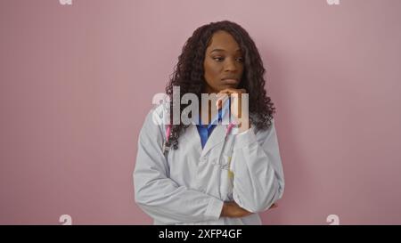 Eine schöne junge afroamerikanische Frau mit lockigem Haar, in medizinischer Kleidung gekleidet, steht nachdenklich vor einem rosa Hintergrund. Stockfoto
