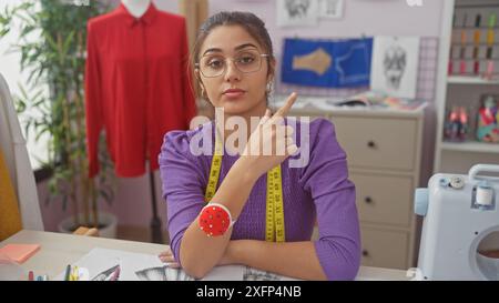 Eine junge hispanische Frau mit Brillenspitzen in einem Schneiderladen, umgeben von Nähgeräten und Kleidungsstücken. Stockfoto