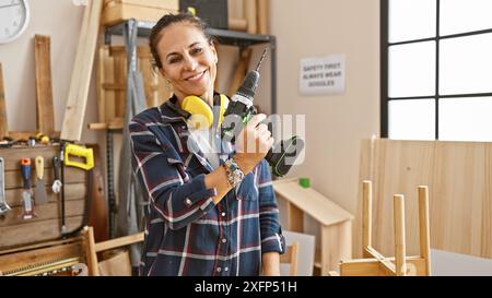 Lächelnde reife hispanische Frau mit lockigem Haar mit Bohrer in sonnigem Holzbearbeitungsstudio. Stockfoto