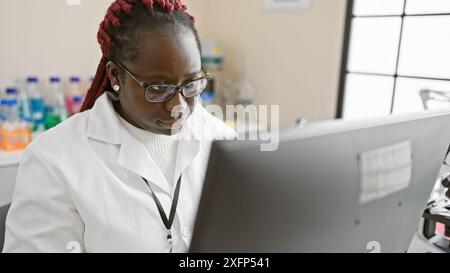 afroamerikanische Wissenschaftlerin mit Zöpfen arbeitet aufmerksam an einem Computer im Labor. Stockfoto