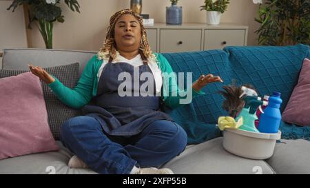 Junge afroamerikanische Frau mit Zöpfen, die zu Hause im Wohnzimmer meditiert, umgeben von Putzmitteln auf der Couch. Stockfoto