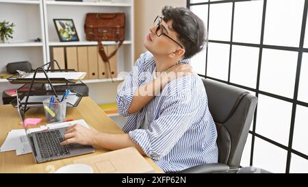 Junger Mann in gestreiftem Hemd, der Nackenschmerzen spürt, während er in einem modernen Büro am Laptop arbeitet. Stockfoto