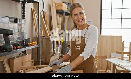 Lächelnde Schreinerin in Schürze, die in einer sonnigen Werkstatt mit Holz arbeitet Stockfoto