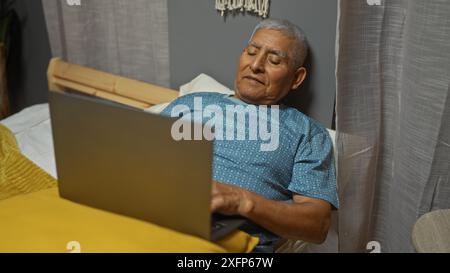 Grauhaariger hispanischer Mann, der in einem Schlafzimmer an einem Laptop arbeitet, auf einem Bett in einer komfortablen Wohnumgebung liegt und eine reife, entspannte Beziehung verkörpert Stockfoto