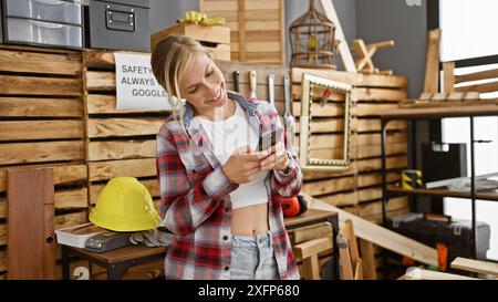 Eine lächelnde junge Frau, die ein Smartphone in einer Tischlerei benutzt, gefüllt mit Werkzeugen und Holzdielen. Stockfoto
