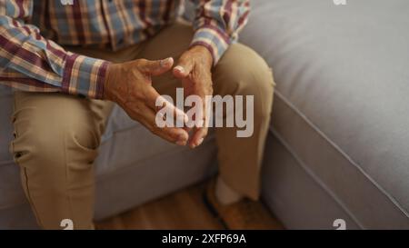 Mann mittleren Alters sitzt drinnen auf einer Couch mit gefalteten Händen, trägt ein kariertes Hemd und eine beige Hose in einer komfortablen Wohnzimmerumgebung. Stockfoto