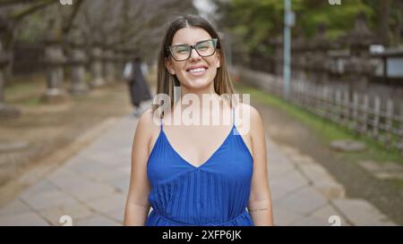 Fröhlicher Ausdruck, schöne hispanische Frau mit Brille, die Positivität und Selbstvertrauen in lässige Kleidung annimmt, während sie strahlend lächelt und zusteht Stockfoto