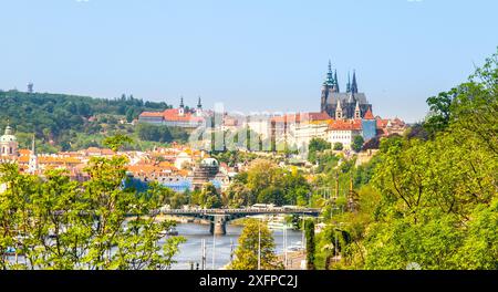 Ein Panoramablick auf die Prager Burg und das Kloster Strahov vom Letna-Garten aus, mit einer Brücke im Vordergrund und üppigen grünen Bäumen, die die Szene umrahmen. Stockfoto