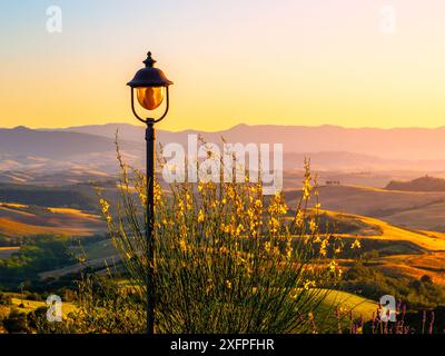 Eine einzige Straßenlaterne steht hoch vor dem Hintergrund der sanften Hügel in der Toskana, Italien. Das warme Leuchten der untergehenden Sonne färbt den Himmel in Orange- und Rosa-Tönen und beleuchtet die Landschaft. Stockfoto