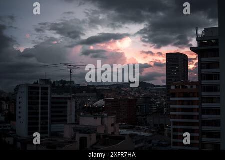 Ein dramatischer Sonnenuntergang überzieht den Himmel in rosa und violetten Tönen über der Stadtlandschaft von Quito, Ecuador, mit Silhouetten von Gebäuden und üppigen Hügeln im Hinterland Stockfoto