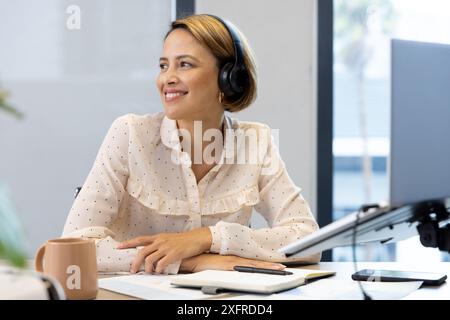 Kopfhörer tragen, Frau lächelt, während sie am Schreibtisch mit Laptop und Notizblock arbeitet Stockfoto