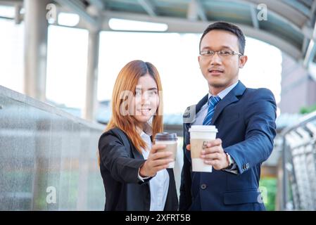 Junge asiatische Geschäftsleute lächeln und entspannen sich bei einer Tasse Kaffee, bevor sie zur Arbeit gehen. Paarliebhaber genießen es, gemeinsam Kaffee in der modernen Stadt zu trinken. Stockfoto