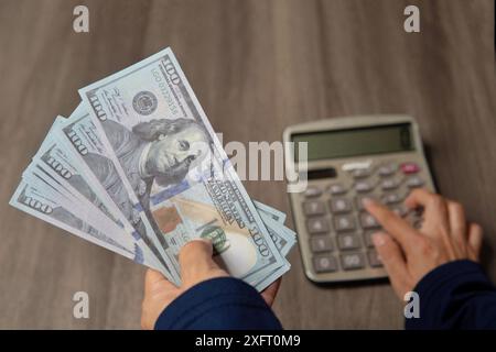 Nahaufnahme der Hände einer Person, die einen Taschenrechner benutzt und einen Haufen Bargeld auf einem Holztisch hält. Finanzkonzept. Stockfoto