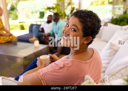 Lächelnde Frau, die eine Tasse Kaffee hält, während sie sich mit verschiedenen Freunden zu Hause trifft Stockfoto
