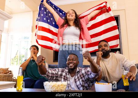 Feiern mit verschiedenen Freunden, Frau mit amerikanischer Flagge, während andere auf der Couch jubeln Stockfoto